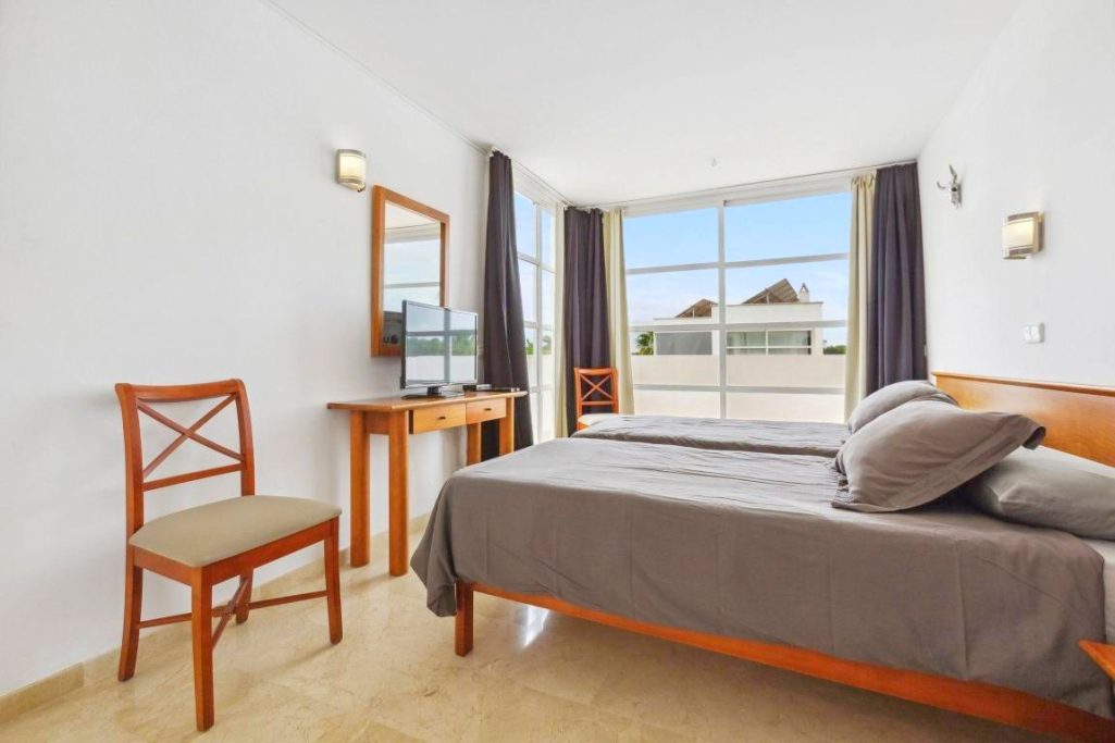 1. Schlafzimmer Helles Zimmer mit zwei Betten, einem Stuhl und einem Tisch, große Fenster mit Sicht nach draußen.