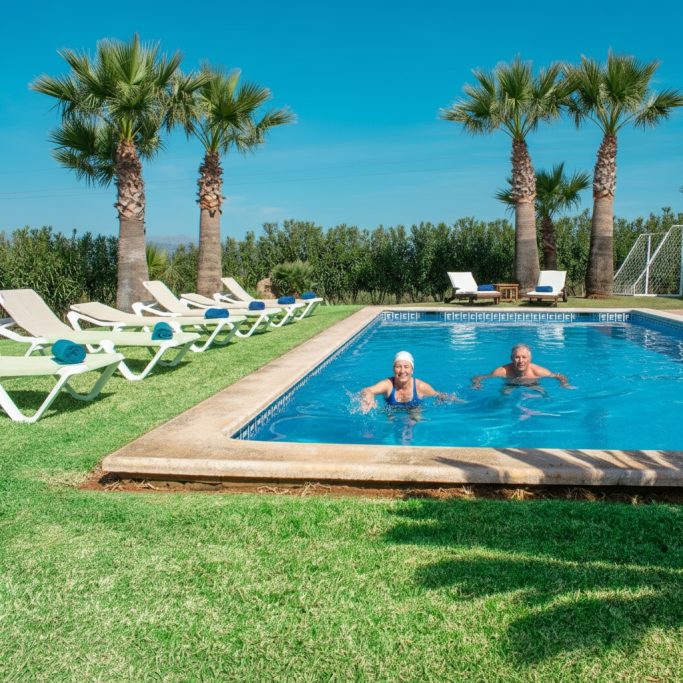 Erfrischung im Pool Zwei Personen schwimmen im Pool umgeben von Palmen und Liegen auf einer grünen Wiese.